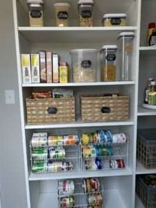 Well organized food storage with containers of pasta and dry goods, with box mixes on the top two shelves, then woven baskets with smaller foods, and canned goods on stackable shelves.