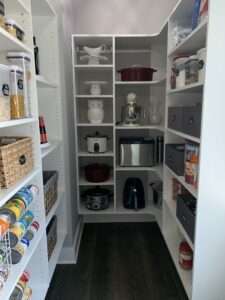 A well-organized kitchen pantry with food in storage containers, woven baskets, and stacked canned goods on the left. And kitchen tools and an white owl cookie jar on the rear wall, and more woven baskets and plastic container food storage on the right side.