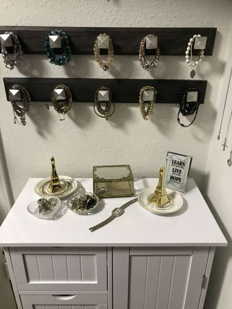 A bracelet holder at the top in two rows above a small white hutch, with 2 golden Eiffel Towers, some bowls with rings, a small silver or pewter box, and a Learn, Live, Hope inscription.