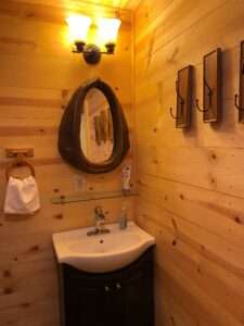 A rustic bathroom, with a mirror set in a piece for a horse, above a sink, with lights above, and hooks framed on the right side. The walls are unfinished, rustic wood.