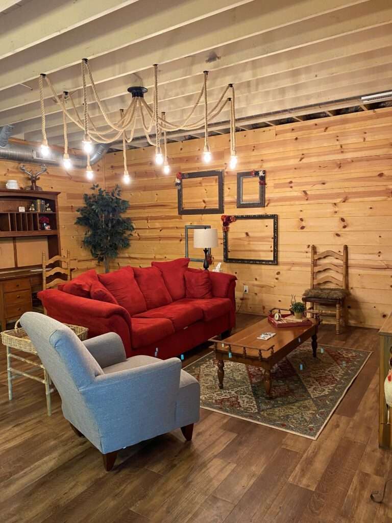 The living room area with a red couch, a gray armchair, coffee table, and 2 wooden straight backed chairs. There is a rope chandelier hanging over the couch, empty black picture frames on the wall, and a rug under the coffee table.