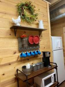 One side of a galley kitchen, with a decorative wreath hanging on the wall, above a shelf with a white ceramic pitcher and white ceramic chicken, above a hanging plate and cup rack with red plates and blue cups, above a standing shelf with the microwave and coffee maker on it. There is a refrigerator to the right of the microwave stand.
