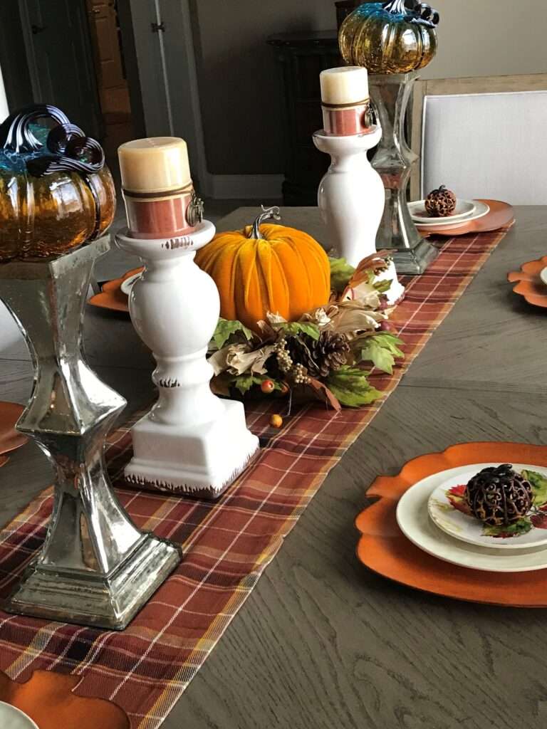 An autumn theme with a red and brown table runner, pumpkin decorations, and 2 candle pedestals holding cream and brown candles.