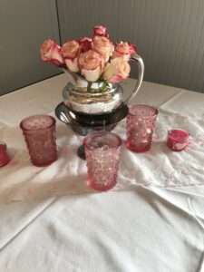 A silver vase with red-tipped pink roses, on a white tablecloth with pink candle holders and candles. There is also a runner going horizontally across the middle third with some pink and white embroidery decoration on it.