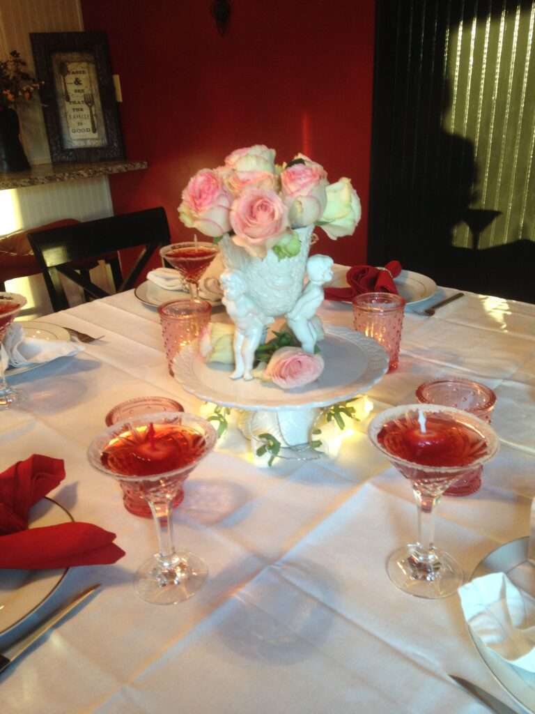 A white tablecloth, white plates, red napkins with string pearl napkin rings, there are candles in red candle holders, and the center piece is a ceramic white glazed piece with 4 cherubs holding a flower vase, with pink tipped white rosses in it.