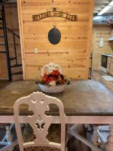 Rustic dining area with a wooden top table with white painted legs and white ornamental chairs, in front of an unfinished wooden wall with 'Welcome to the Barn' in metal work.
