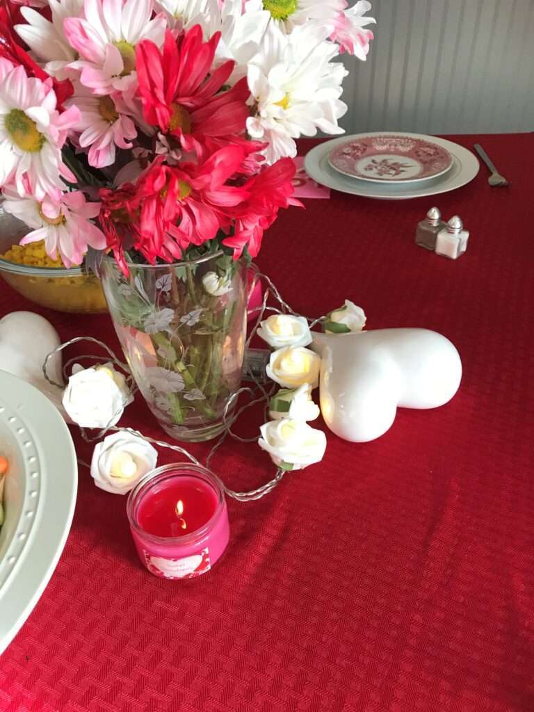 A close-up of a table center piece of red, white, and pink flowers, in a clear vase, on a red tablecloth, with a 3-D white heart, and some flower light decorations and pink candles. The place settings have a red and white motif.