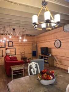 Living room and dining area in this rustic Airbnb. Empty black photo frames hang on the wall in the back left, some blankets hang in the corner on a ladder, and a tv and clock are on the right wall. There is a wooden chair, red couch, and gray armchair. Up close is the dark brown wooden table, with a floral arrangement of reds and oranges in a distressed white wash basin. There are white backed ornamental chairs.