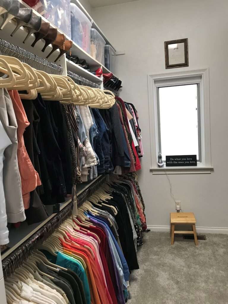 A closet wall, with plastic bins at the top, shoes along the middle and two rows of pants and shirts at the bottom of the wall.