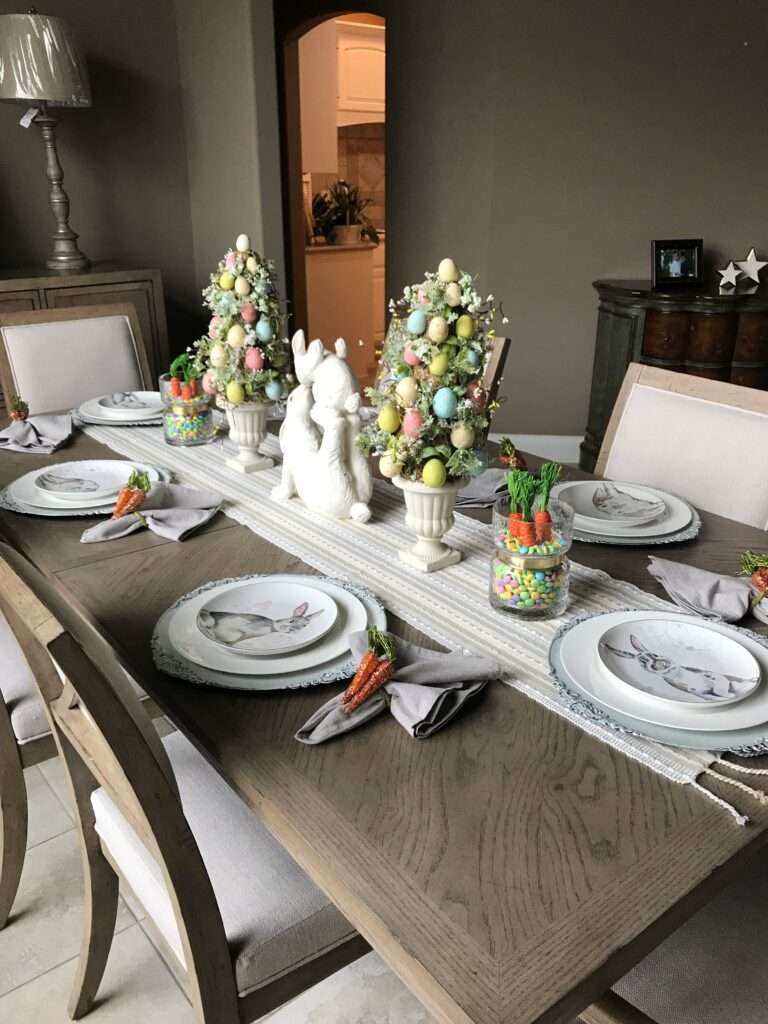 An Easter tablescape with egg "trees" in white plant bases on either side of a white ceramic bunny rabbit on his back, feet in the air, playing with an egg. On the furthest out from the bunny, there are carrots in jars with candy. The napkin rings are also carrots. And the plates have pictures of a bunny on them.