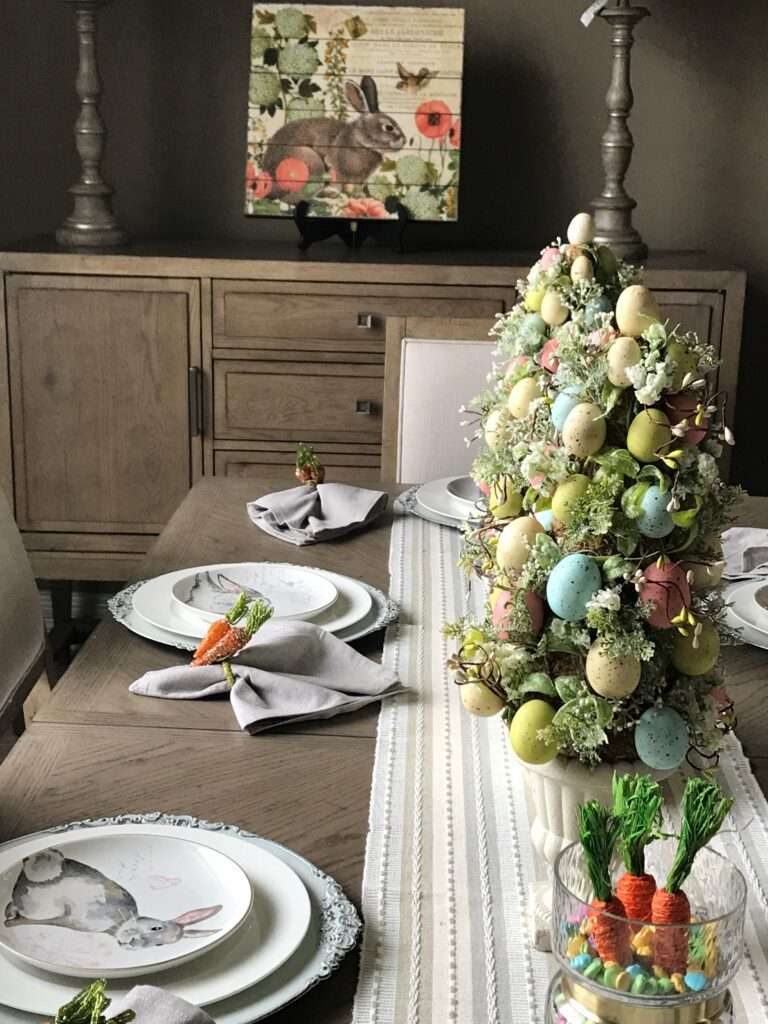 An Easter tablescape with egg "trees" in white plant bases on either side of a white ceramic bunny rabbit on his back, feet in the air, playing with an egg. On the furthest out from the bunny, there are carrots in jars with candy. The napkin rings are also carrots. And the plates have pictures of a bunny on them. On the back wall is a wall hanging with a rabbit, and some pink and green flowers.