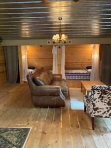 Seating area and bed nooks. There is a dark leather couch, a multicolored leather chair, and a wooden coffee table over a white rug. In the background are two bed nooks with blue blankets, blue accent pillows, and shear curtains to draw them closed.