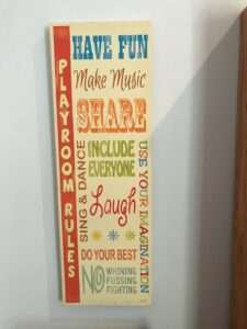 A sign: Playroom Rules: Have fun, Make Music, Share, Include Everyone, Sing and Dance, Laugh, Use Your Imagination, Do Your Best, No Whining, Fussing, or Fighting.