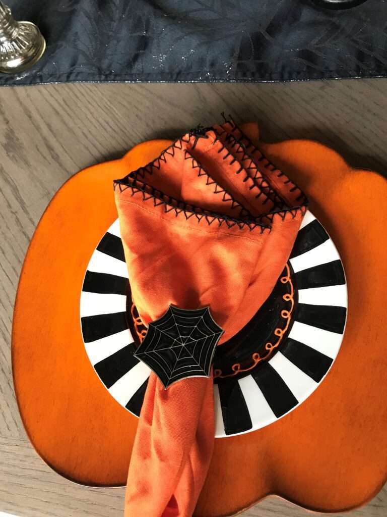 Table setting with orange napkins, pumpkin shaped table cloth, and black and white striped plates. The napkin ring is a spider web.