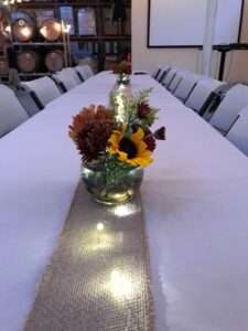 A cream colored table cloth with a burlap strip down the middle fifth, and on the burlap, are a strip of fairy lights with small glass clobe vases with red and orange flowers and some greenery.
