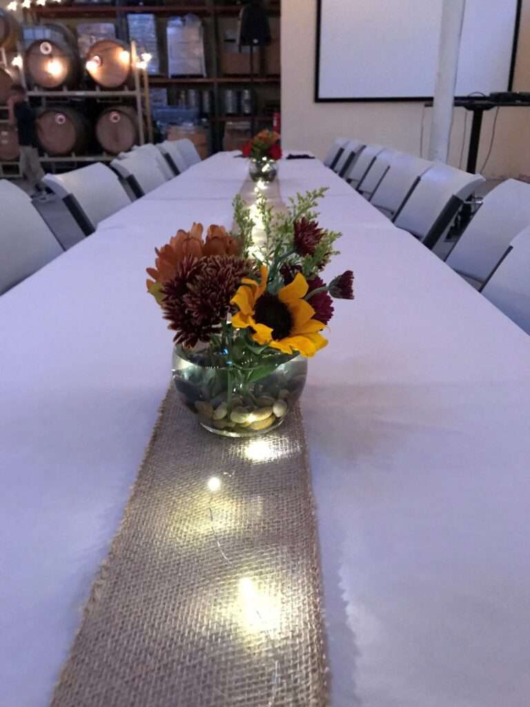 A cream colored table cloth with a burlap strip down the middle fifth, and on the burlap, are a strip of fairy lights with small glass clobe vases with red and orange flowers and some greenery.