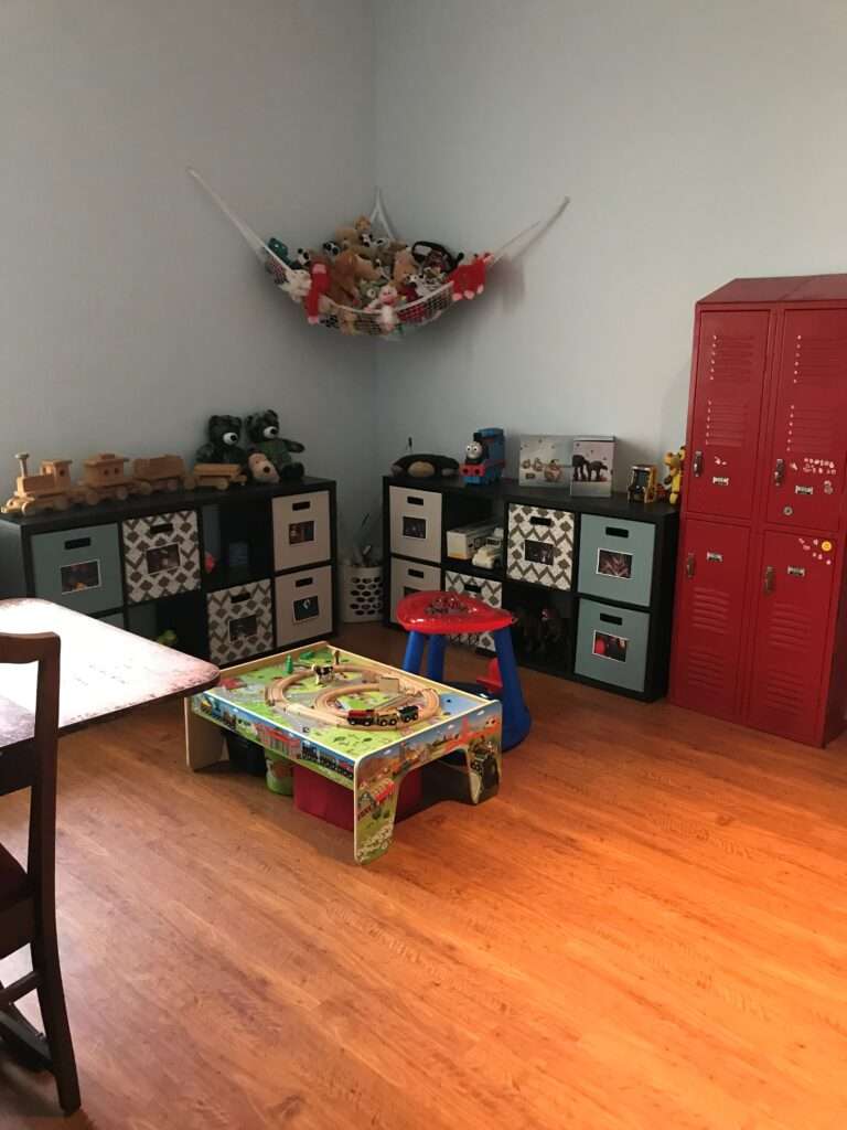 An animal hammock hanging on the wall, and cube bin storage on both side of the corner, on the viewer's right is a 2x2 set of red storage lockers. There is a small play table on the floor.