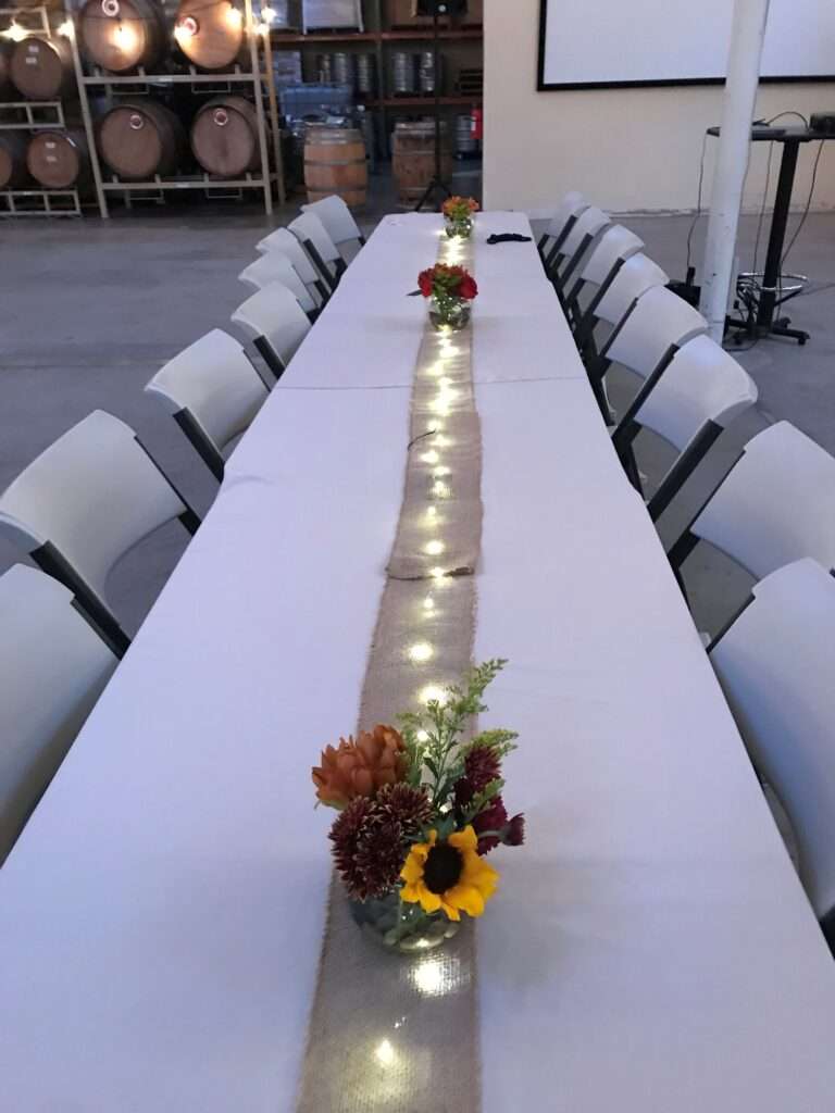 A cream colored table cloth with a burlap strip down the middle fifth, and on the burlap, are a strip of fairy lights with small glass clobe vases with red and orange flowers and some greenery.
