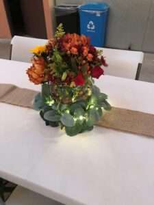 A cream colored table cloth with a burlap strip down the middle fifth, and on the burlap, are a strip of fairy lights with small glass clobe vases with red and orange flowers and some greenery.