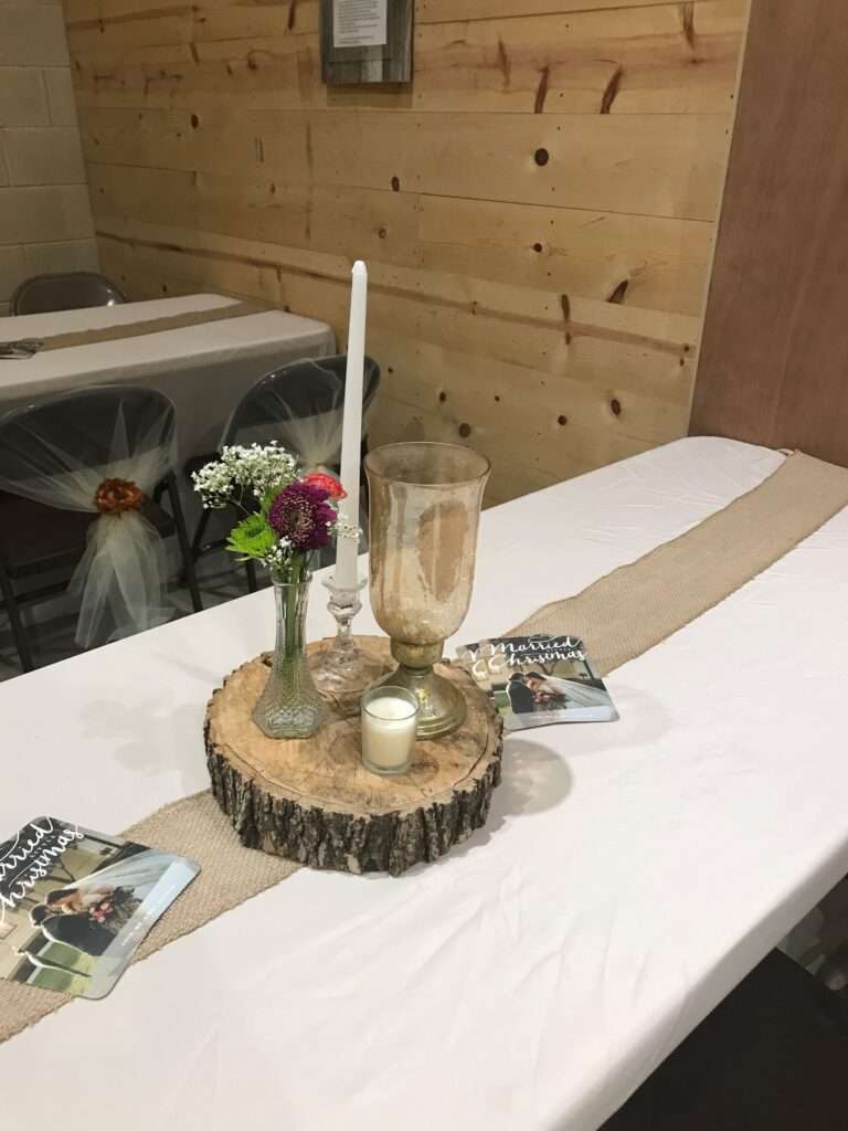 A piece of log with a tall glass vase with red, pink, and white flowers, and white candle in a glass candle holder, and an empty amber colored glass vase and a small white candle in a jar.