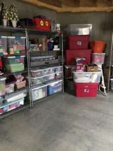 2 tall metal shelves in a garage, filled with totes of organized holiday decorations. There are some decorations stacked to the side.