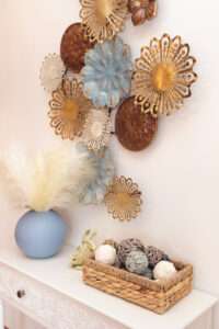Close up of natural décor in a basket on a white table. A blue and gold wall hanging hangs over it.