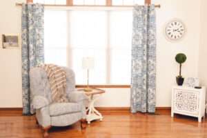 Gray-blue chair in front a window with blue and white curtains.