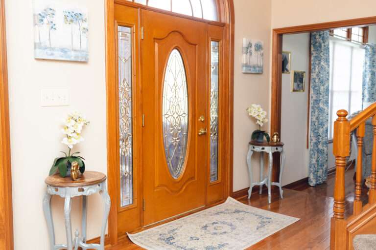Front door flanked by small circular tables with silvery-gray legs and wooden tops, each adorned with white orchids and gold frogs. Above each table is an image of blue and gold trees over a white background. There is also a blue and white carpet in front of the door.