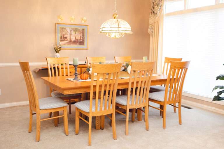 A wooden table, with a fall pumpkin themed runner, and a large central floral arrangement with greenery, blue and white flowers, at either end at pewter candle holders with moss green candles. There are small decorative pumpkins next to the candle holders. This picture shows the entire table, with 8 chairs.