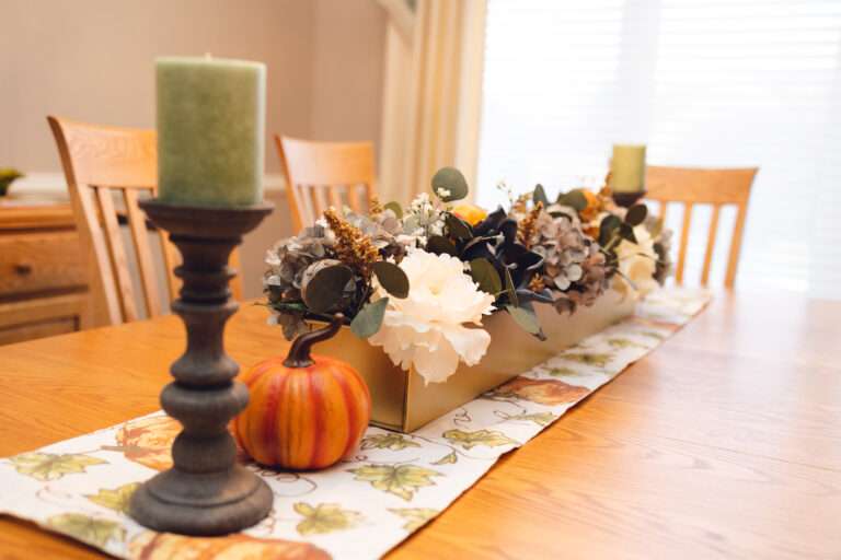 A wooden table, with a fall pumpkin themed runner, and a large central floral arrangement with greenery, blue and white flowers, at either end at pewter candle holders with moss green candles. There are small decorative pumpkins next to the candle holders.