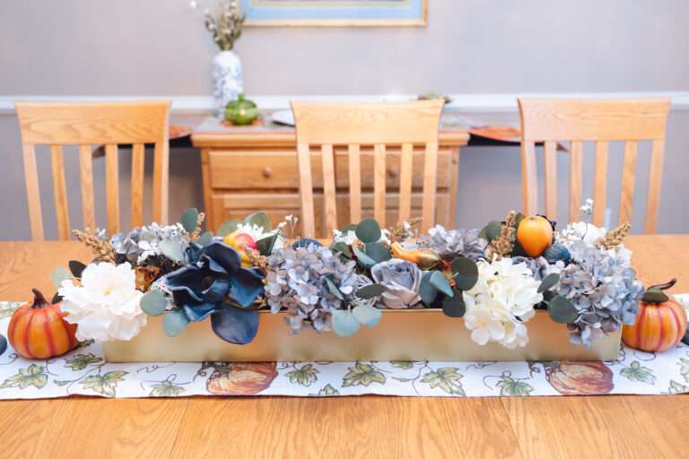 A wooden table, with a fall pumpkin themed runner, and a large central floral arrangement with greenery, blue and white flowers, at either end are small decorative pumpkins.