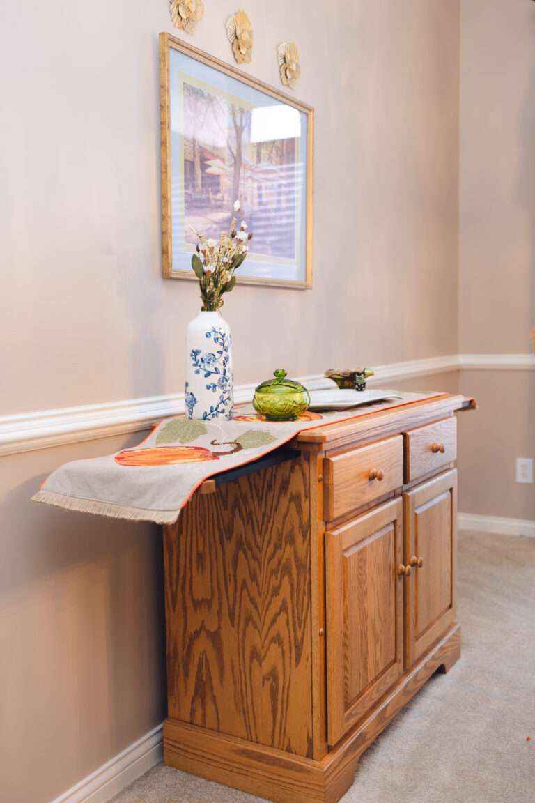 A side table with a blue and white floral vase, a small green glass dish, and white serving tray with white pear handles. There is a fall pumpkin themed runner and the table is extended for serving.