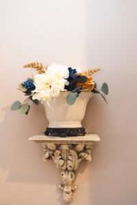 A close up of a decorative wall sconce. The sconce is very ornamental with leaves in an antique white. There is a white planter with dark feet on top, holding a floral arrangement with green leaves, white, blue, and orange flowers, and some feathered tan accent plants.