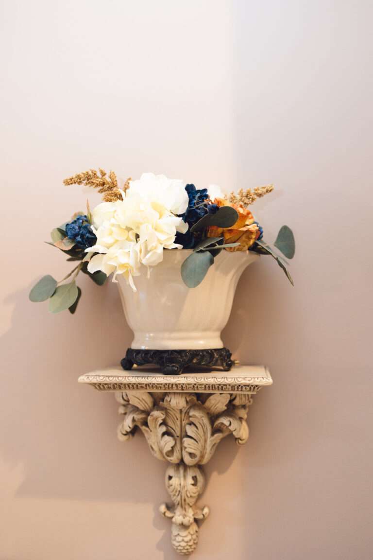 A close up of a decorative wall sconce. The sconce is very ornamental with leaves in an antique white. There is a white planter with dark feet on top, holding a floral arrangement with green leaves, white, blue, and orange flowers, and some feathered tan accent plants.
