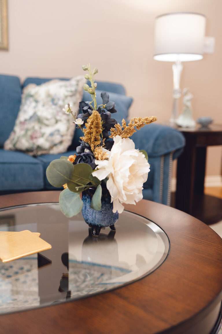 A close up of a small blue vase on a glass topped wooden table, holding blue and white flowers, with green leaves, and tan floral accents.