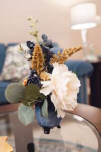 A close up of a small blue vase on a glass topped wooden table, holding blue and white flowers, with green leaves, and tan floral accents.
