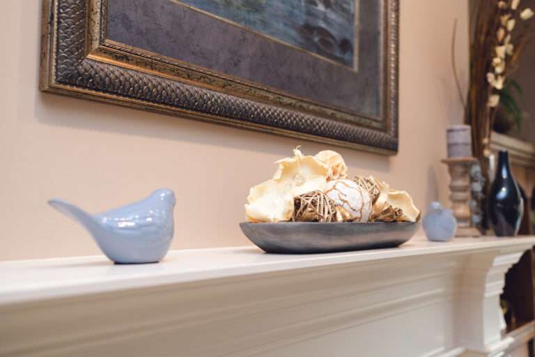 Fireplace mantel with small blue ceramic birds, a blue image in a antique silvery frame, with accent stone colored candle holders, and shiny dark slate colors vases, and a clear vase with tall grass-like floral arrangement with small white flowers.