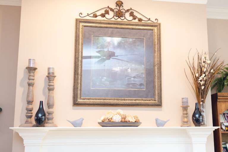 Fireplace mantel with small blue ceramic birds, a blue image in a antique silvery frame, with accent stone colored candle holders, and shiny dark slate colors vases, and a clear vase with tall grass-like floral arrangement with small white flowers.
