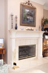 Fireplace mantel with small blue ceramic birds, a blue image in a antique silvery frame, with accent stone colored candle holders, and shiny dark slate colors vases, and a clear vase with tall grass-like floral arrangement with small white flowers. The while tiled fireplace is visible in this picture.