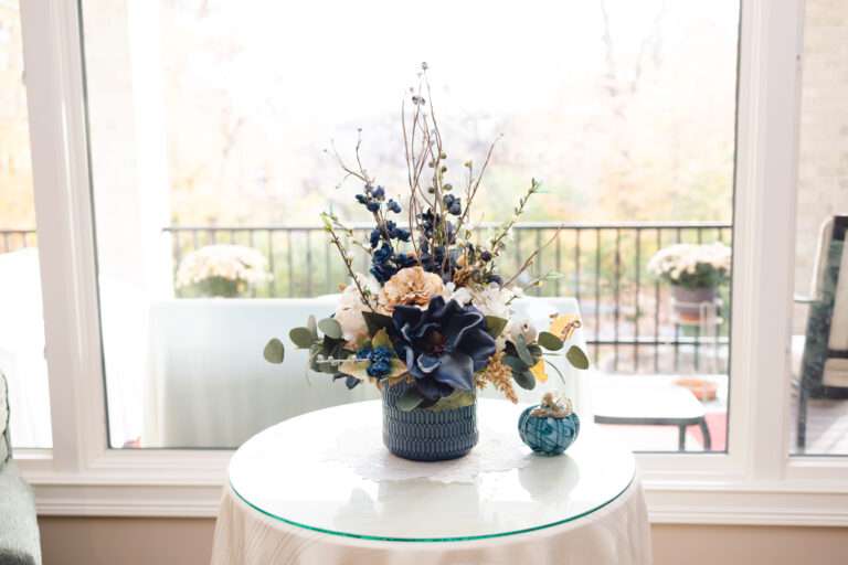 Wise shot of a blue and white floral arrangement on a small table with a white table cloth and glass top.