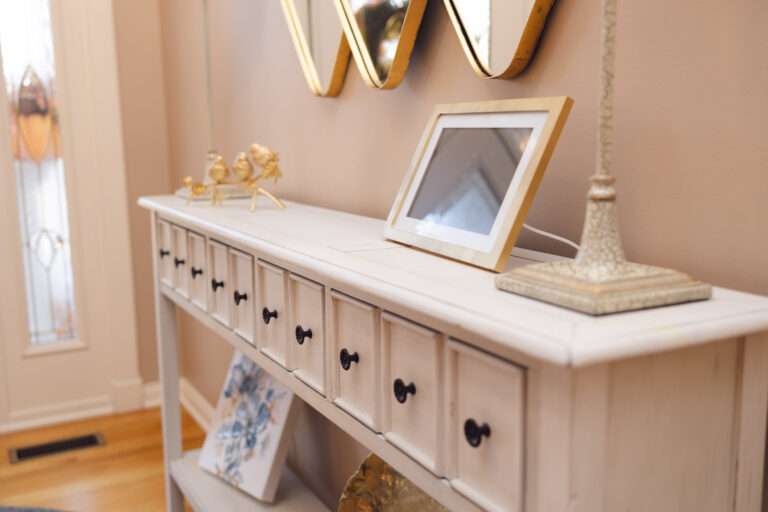 A small white console table with lots of drawers, a gold bird statue, picture frame, some blue, white, and gold accent pieces, under a gold edged mirror hanging on the wall.