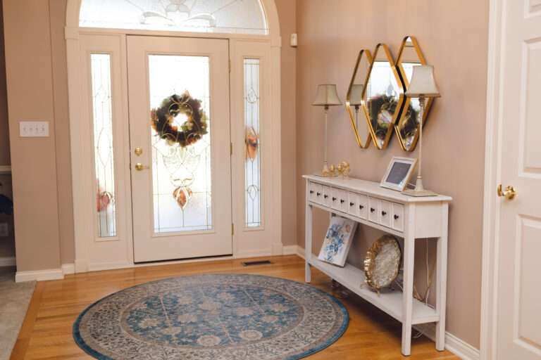 A small white console table with lots of drawers, a gold bird statue, picture frame, some blue, white, and gold accent pieces, under a gold edged mirror hanging on the wall. The table is sitting in an entry way, showing the white antiqued lamps on top, and the mirror above. There is a blue distressed rug in front of a white glass door.