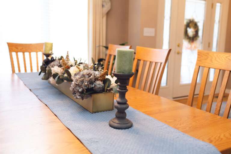 A wooden table, with a blue runner, and a large central floral arrangement with greenery, blue and white flowers, at either end at pewter candle holders with moss green candles.