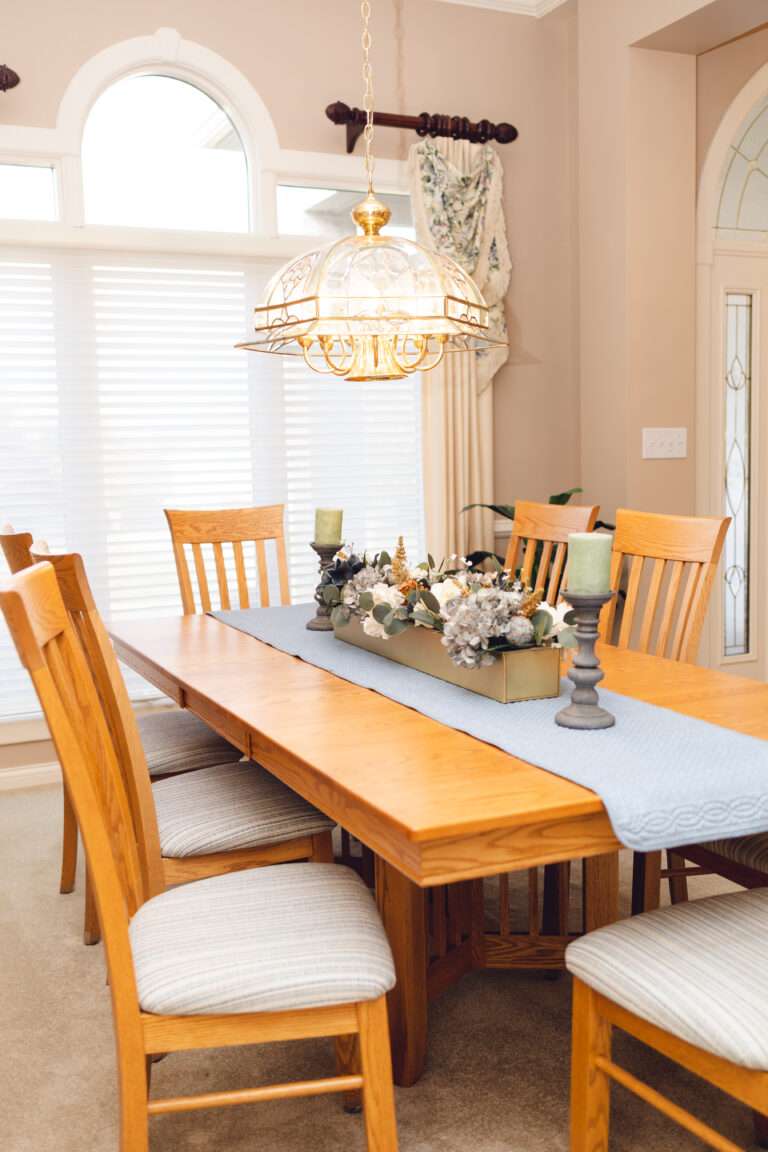 A wooden table, with a blue runner, and a large central floral arrangement with greenery, blue and white flowers, at either end at pewter candle holders with moss green candles.