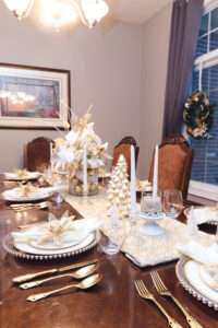 Gold and white Christmas tablescape, with white runner, white and gold Christmas tree, and floral arrangement with white and gold ornaments over a rich brown wooden table.