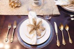 A napkin with a gold and white flower arranged on a white plate, above a clear plate with decorative gold edging, surrounded by gold flatware.