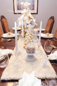 Gold and white Christmas tablescape, with white runner, white and gold Christmas tree, and floral arrangement with white and gold ornaments over a rich brown wooden table.