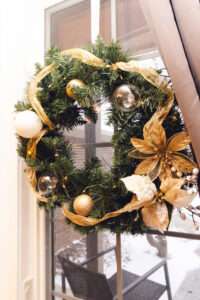 A green wreath with a gold ribbon running around, and gold, clear, and white ornaments.