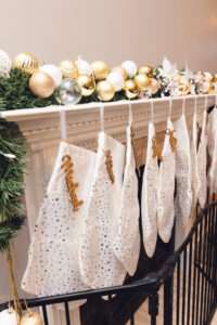 A mantle with some greenery, an abundance of gold, white, and clear ornaments, and many white stockings hung with gold decorations.