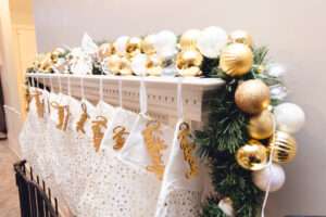 A mantle with some greenery, an abundance of gold, white, and clear ornaments, and many white stockings hung with gold decorations.